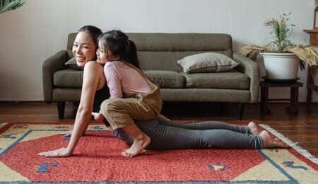 mulher praticando alongamento na sala para evitar os tipos de depressão com sua filha segurando nas costas da mãe 