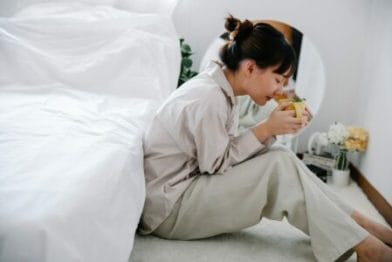 mulher sentada ao lado da cama tomando chá como uma das formas baratas para aliviar o estresse